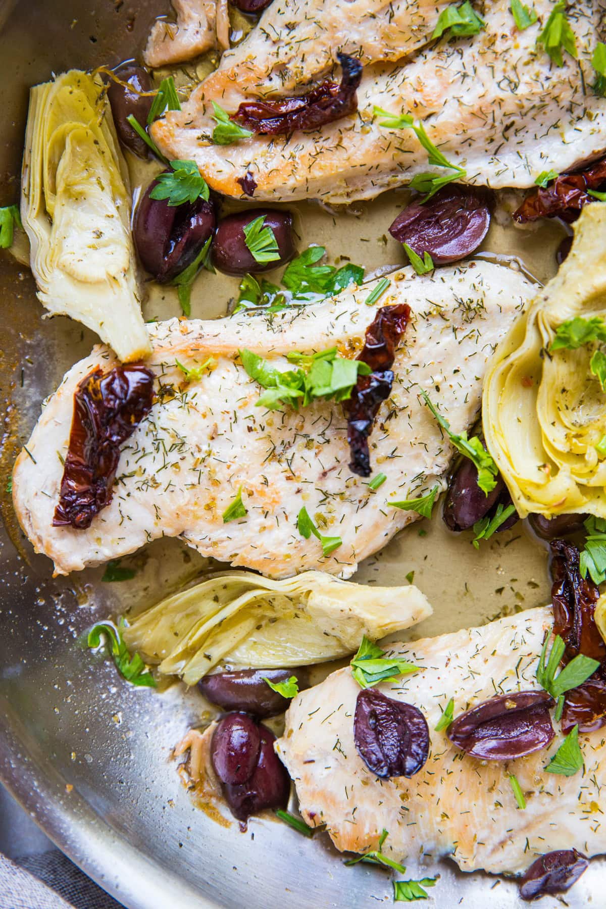 Stainless steel skillet with chicken breasts, sun-dried tomatoes, kalamata olives, and artichoke hearts.