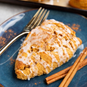 Pumpkin scone on a blue plate covered with glaze and sprinkled with pumpkin pie spice with cinnamon sticks to the side and a baking sheet of scones in the background.