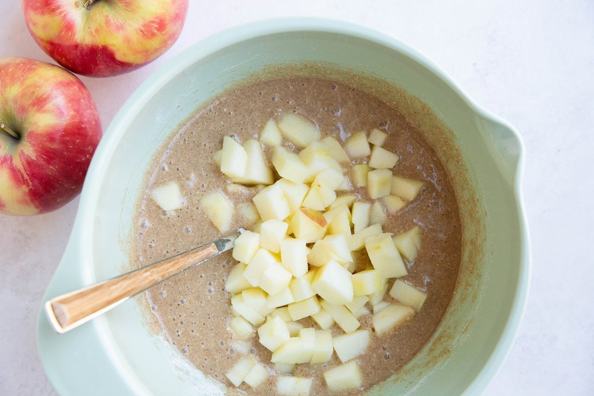 Mixing bowl of apple cake batter with fresh apples on top, ready to be mixed in.