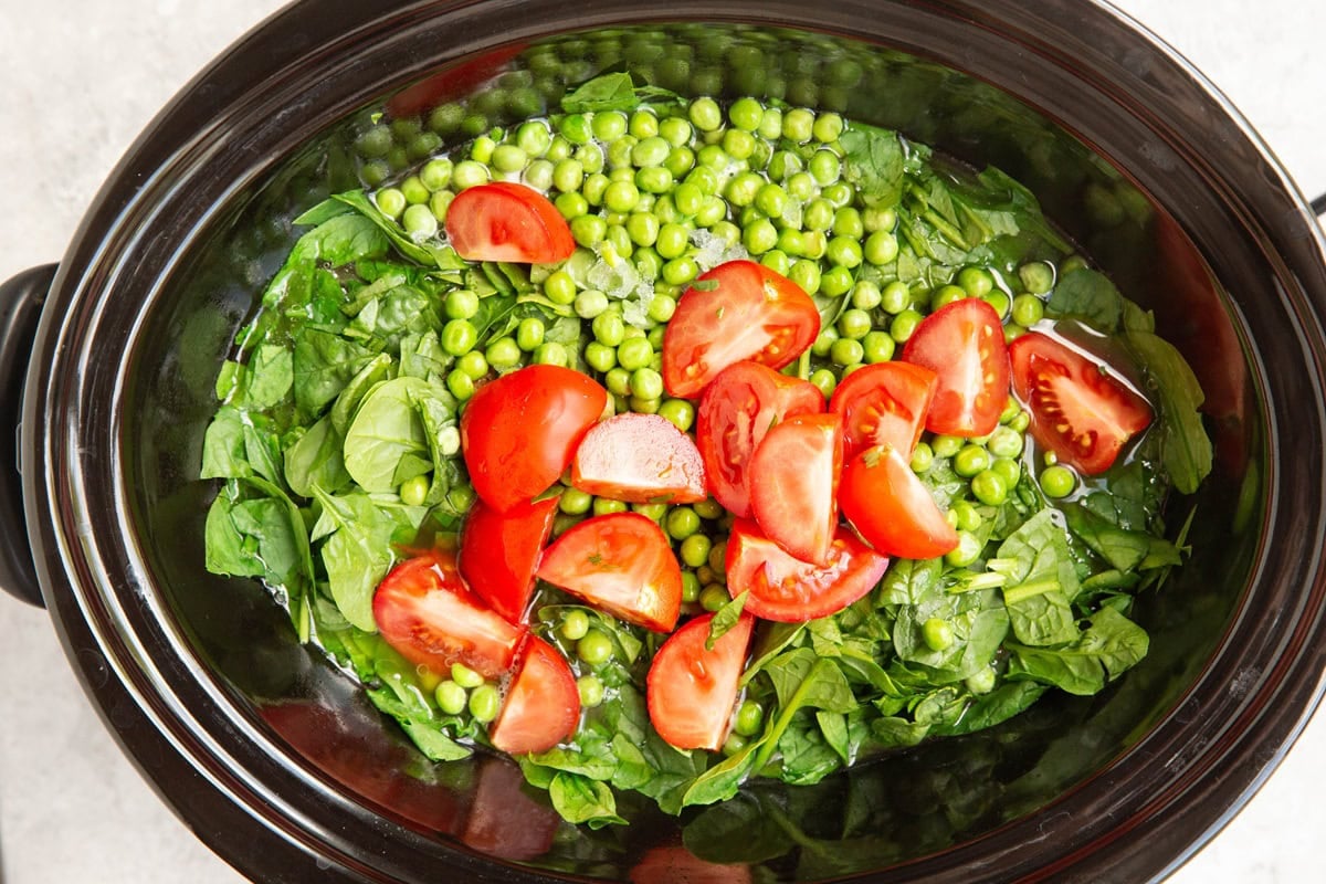 Slow cooker with chicken stew on the bottom, and chopped spinach, peas, and tomatoes on top, ready to be cooked in.