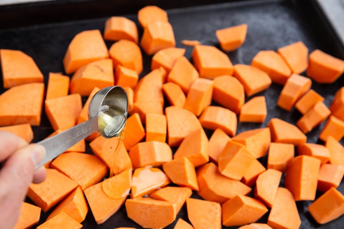 Hand pouring avocado oil over chunks of sweet potato laid out on a baking sheet.