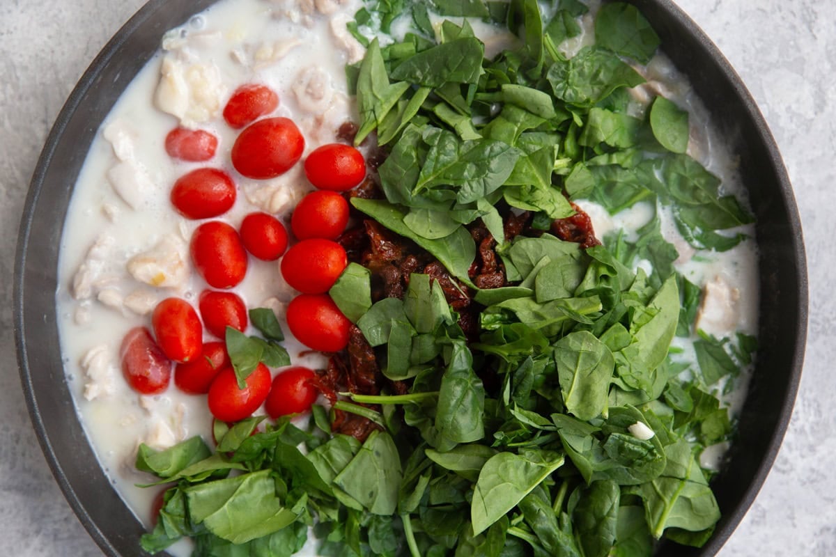 Large skillet full of chicken, onion, coconut milk, sun-dried tomatoes, cherry tomatoes, and baby spinach.