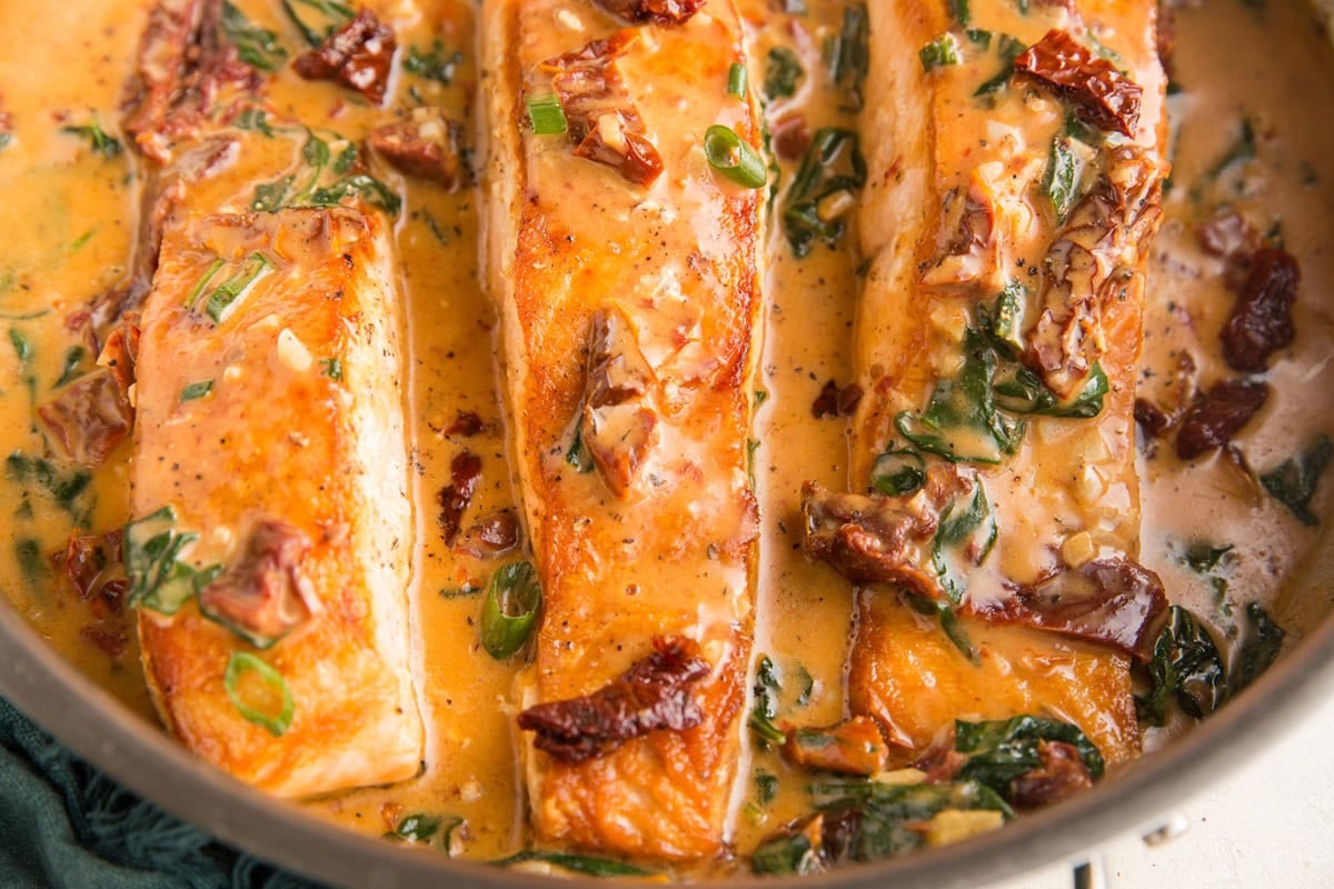 Stainless steel skillet with three salmon filets covered in a creamy sun-dried tomato sauce.
