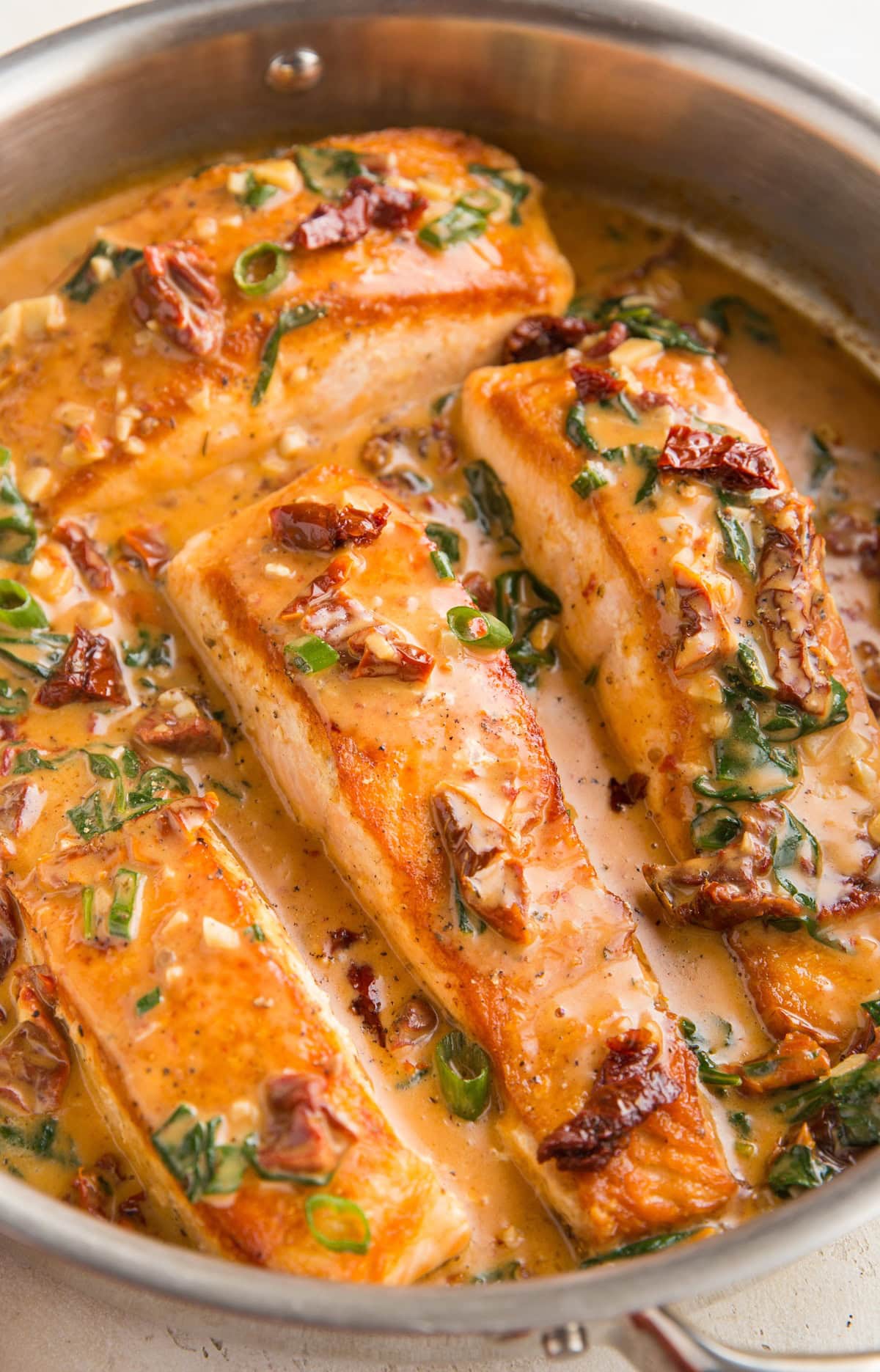 Four cooked salmon filets in a stainless steel skillet with creamy sun-dried tomato sauce. Ready to serve.