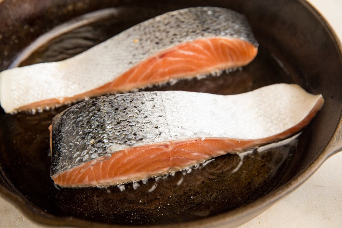 Two salmon fillets being seared skin side up in a cast iron skillet