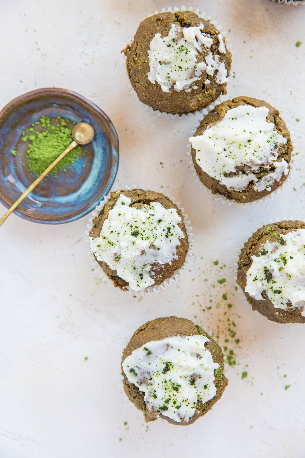 Five matcha muffins sitting on a white background with a small bowl of matcha with a spoon to the side.
