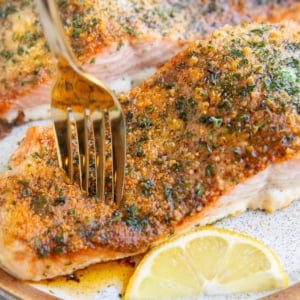 Two parmesan crusted salmon filets on a plate with a fork taking a bite and a lemon wedge to the side.