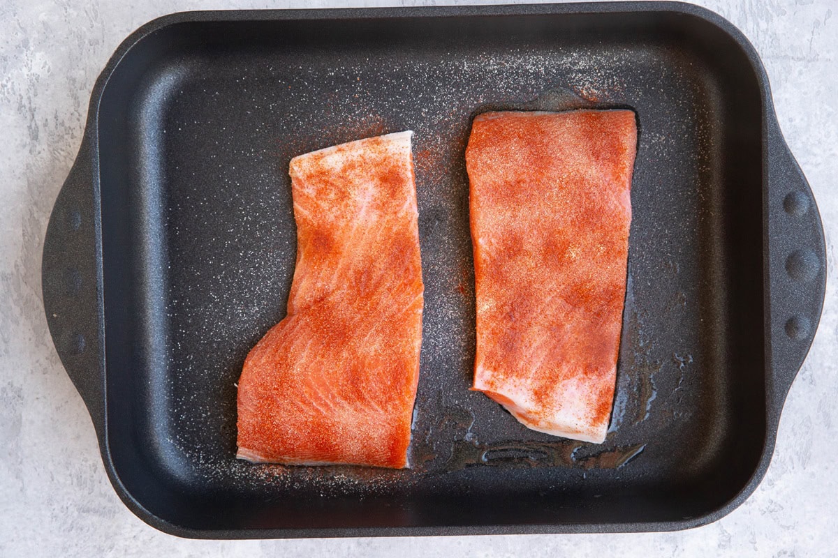 Two raw salmon filets in a casserole dish sprinkled with seasonings.