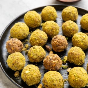 Pistachio energy balls on a black plate with chopped pistachios sprinkled all around. A wooden bowl of ground up pistachios in the background and a golden napkin.