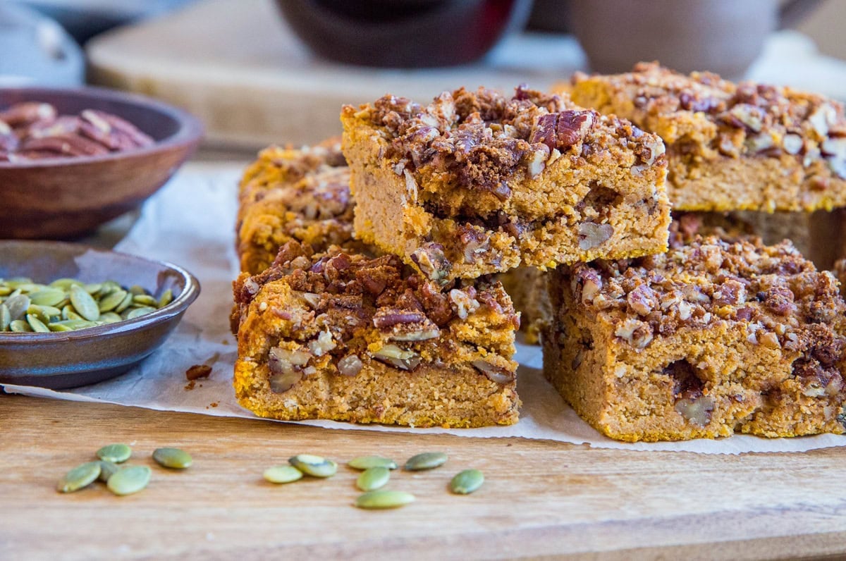 Wooden cutting board with slices of pumpkin coffee cake on top with bowls of pumpkin seeds and pecans in the background.