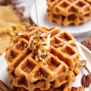 Drizzling honey over three sweet potato waffles with butter and chopped pecans on top. A plate of two waffles in the backfround.