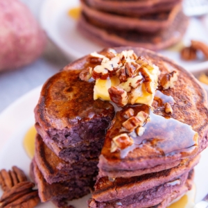 Stack of purple pancakes on a white plate with a bite taken out, topped with butter and chopped pecans.