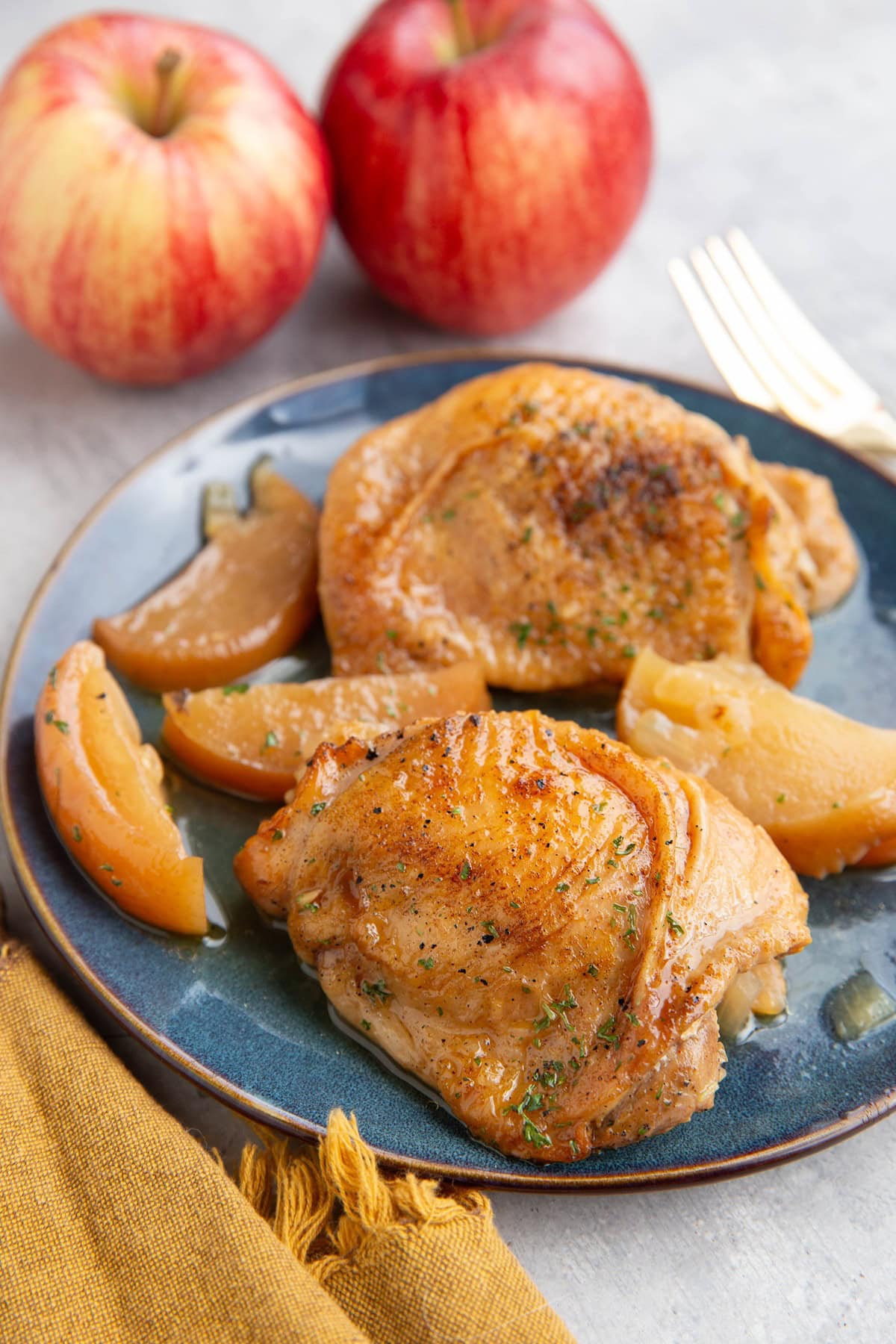 Cooked chicken thighs on a blue plate with cooked apples and fresh apples in the background.