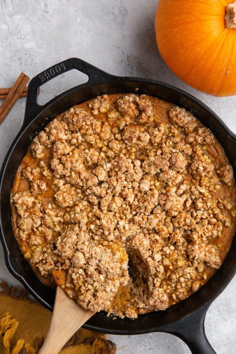 Cast iron skillet full of pumpkin crisp with a wooden spoon scooping out some of the crisp and a fresh pumpkin to the side.