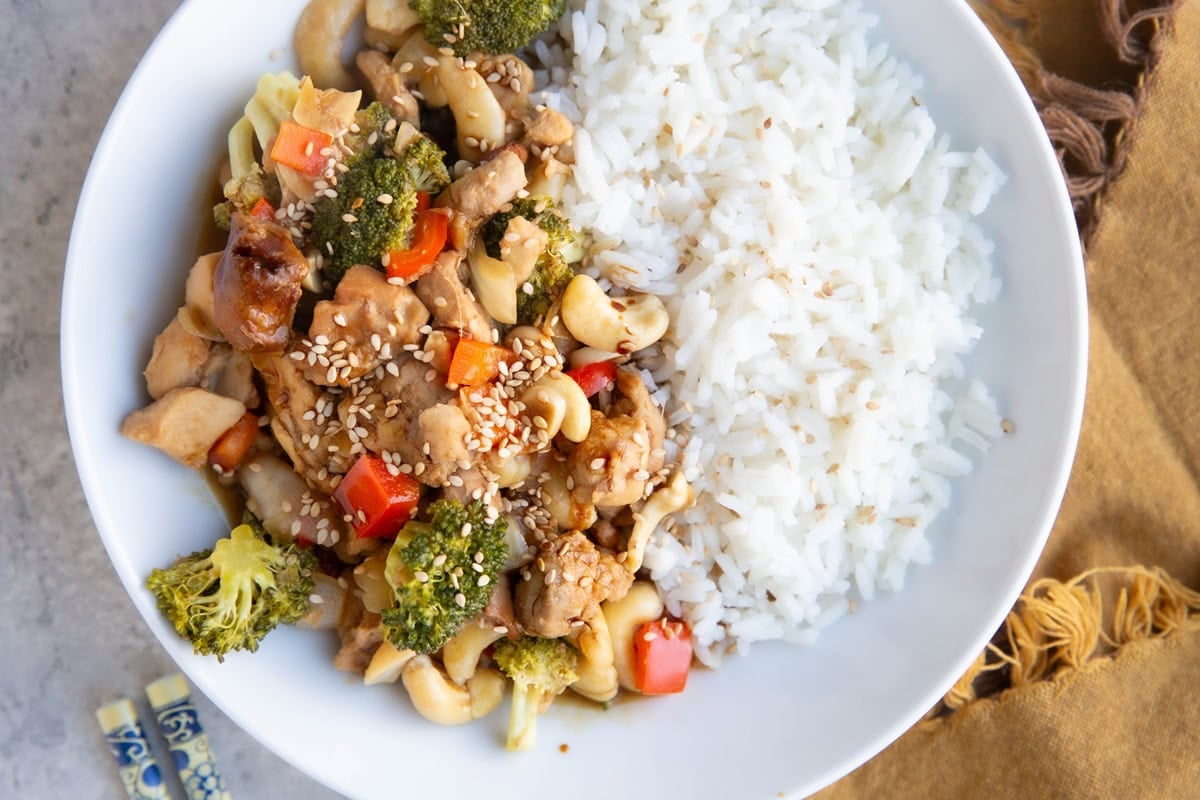 Cashew chicken and rice in a white bowl. Golden napkin and chopsticks to the side.