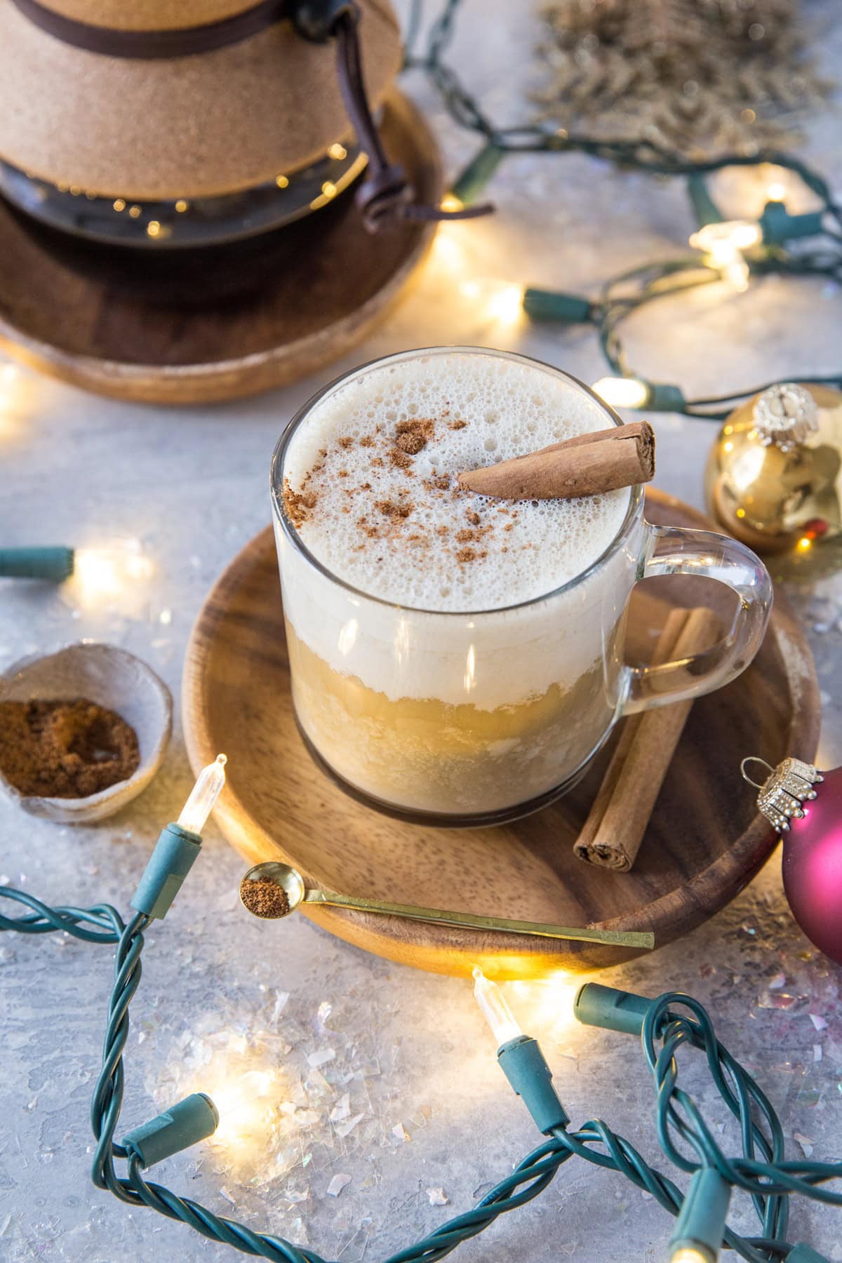 Clear mug full of eggnog latte with Christmas lights and ornaments surrounding the latte with a vat of coffee in the background.