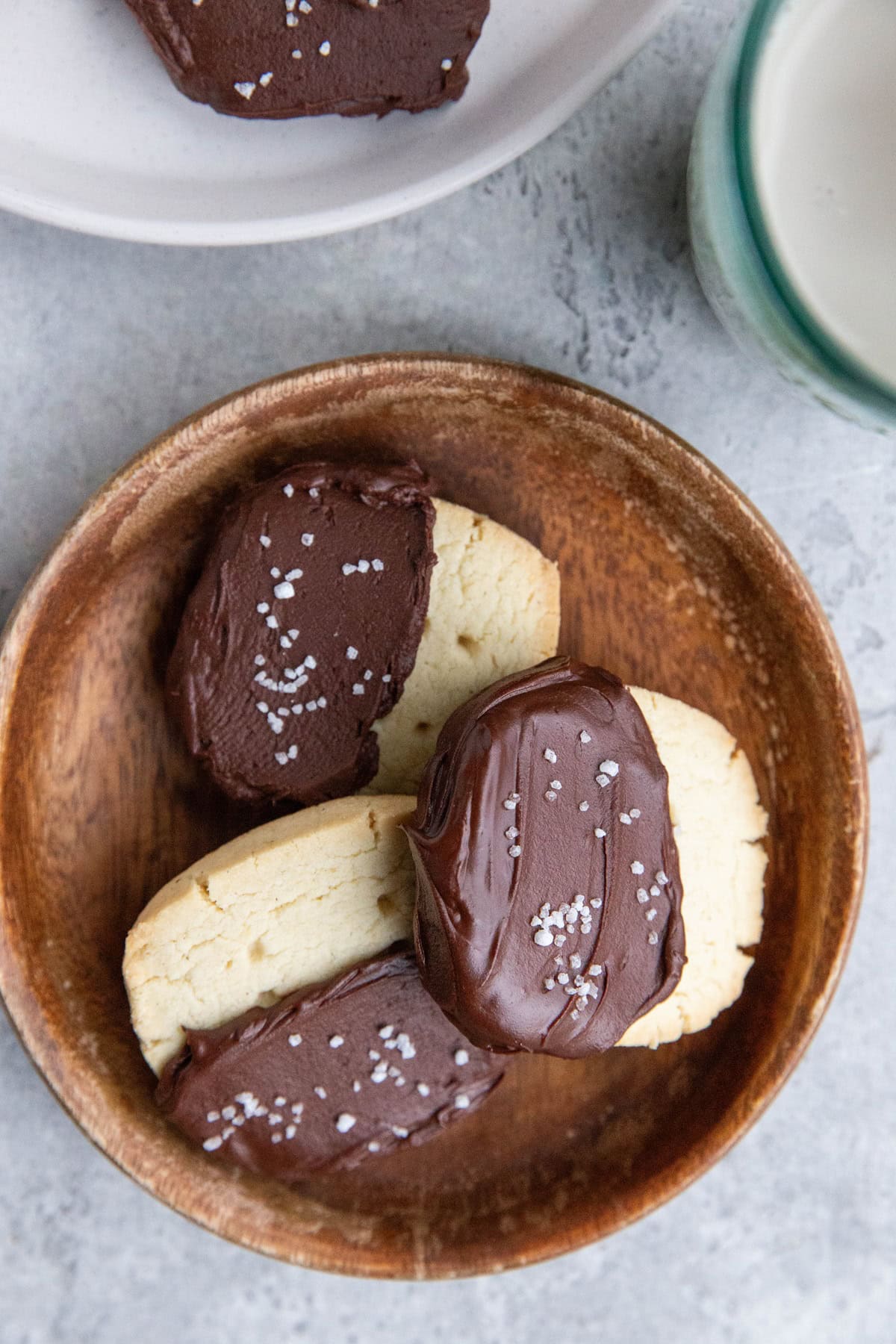 Wooden plate with three shortbread cookies dipped in chocolate. Another plate and a glass of milk to the side.