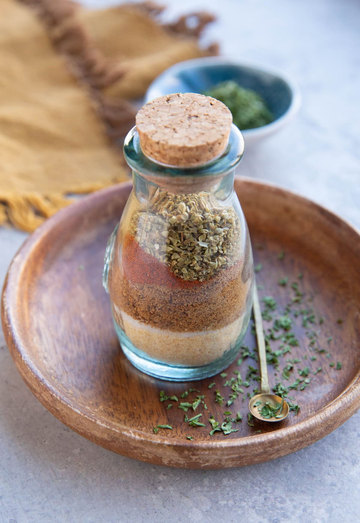 prime rub seasoning blend inside of a small glass jar with a cork sealing the top sitting on a wooden plate with dried parsley all around.