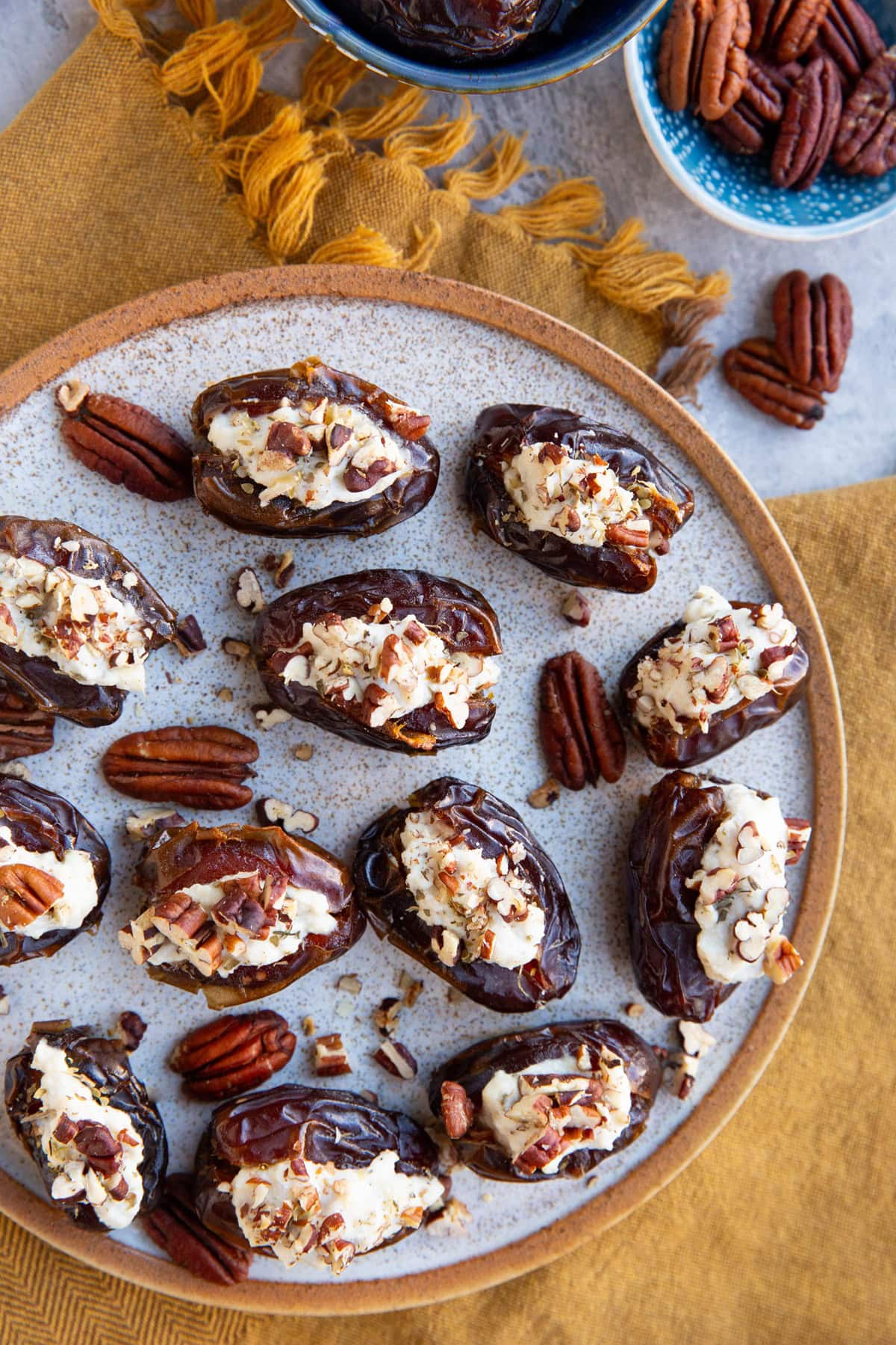 Ricotta stuffed dates on a plate sprinkled with chopped pecans sitting on top of a golden napkin. A bowl of pecans to the side.