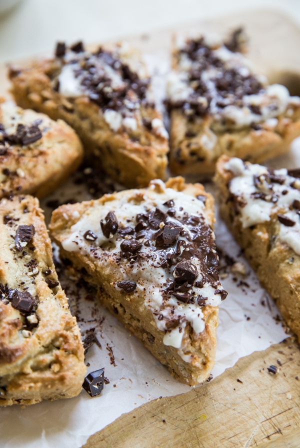 Eight scones sitting on a wooden cutting board that have a white glaze on top and are topped with chopped dark chocolate. Ready to serve.