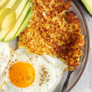 Cauliflower hash browns on a plate with sunny side up eggs and sliced avocado with a fork to the side.