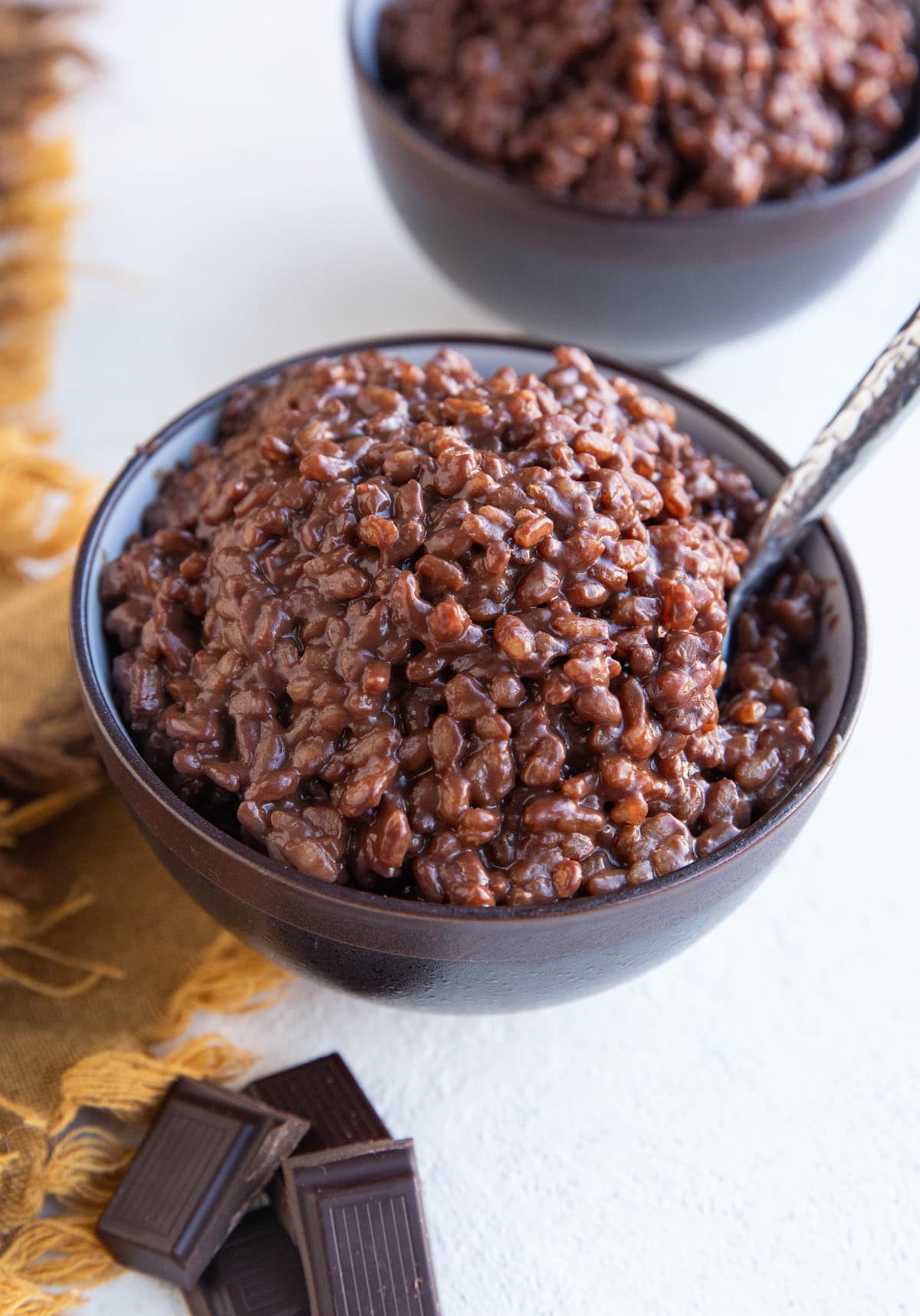 Two bowls full of chocolate rice pudding with chunks of dark chocolate next to one of the bowls and a golden napkin to the side, ready to serve.