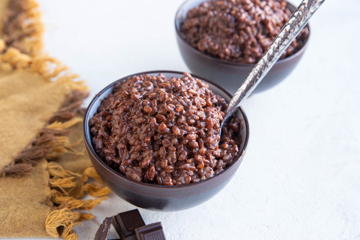 Two bowls of chocolate rice pudding with a spoon and chunks of dark chocolate and a golden napkin.