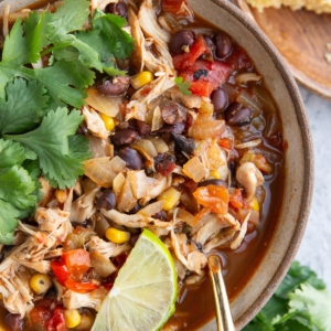 Large bowl full of shredded chicken chili with black beans and corn with fresh cilantro and a slice of lime on top. A plate of cornbread with butter and honey to the side.