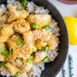 large bowl full of brown rice and lemon chicken with green onions and sesame seeds on top and chop sticks to the side. A wooden bowl sits to the side with chopped green onions.