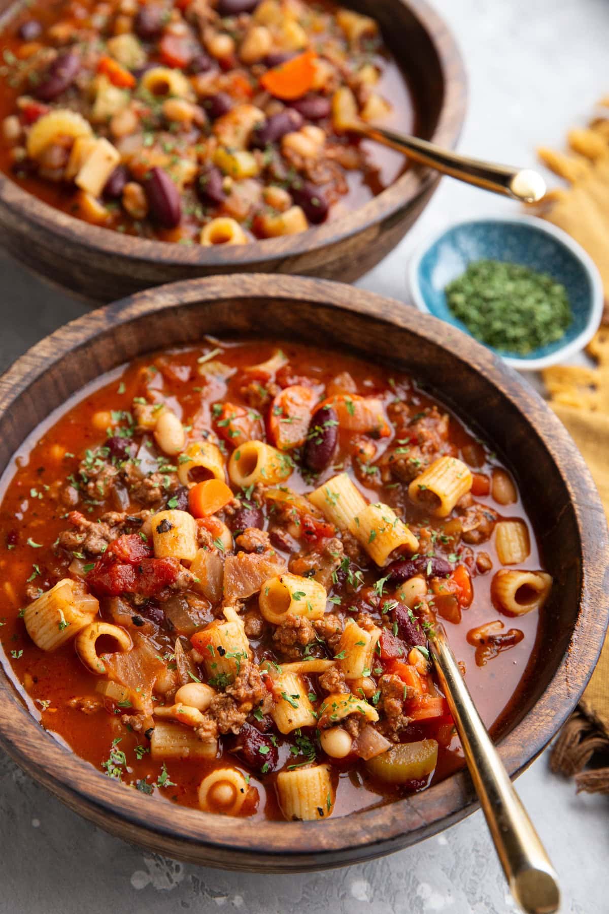 Wooden bowls full of pasta e fagioli soup with a small bowl of dried parsley in the background and golden spoons inside of the soup bowls, ready to eat.