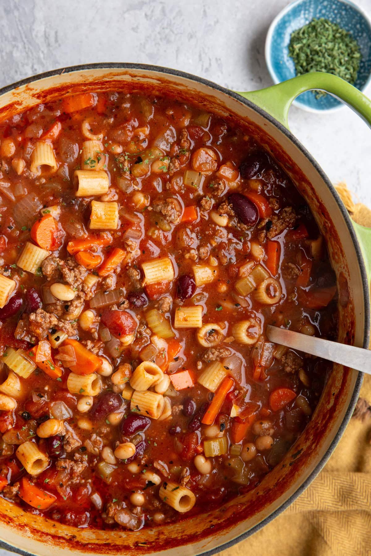 Large soup pot full of Italian pasta soup with beans and beef and a small blue bowl of dried parsley to the side.