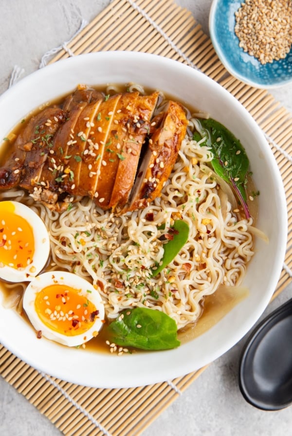 Big white bowl full of ramen noodles with broth, sliced chicken and soft boiled egg. Sprinkled with sesame seeds, red pepper flakes, and parsley with a black soup spoon to the side and a small blue bowl of sesame seeds.