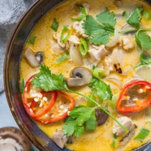 Blue ceramic bowl full of creamy Thai coconut chicken soup with mushrooms, cilantro, and red chilis on top. A fresh mushroom and half of a lime to the side.