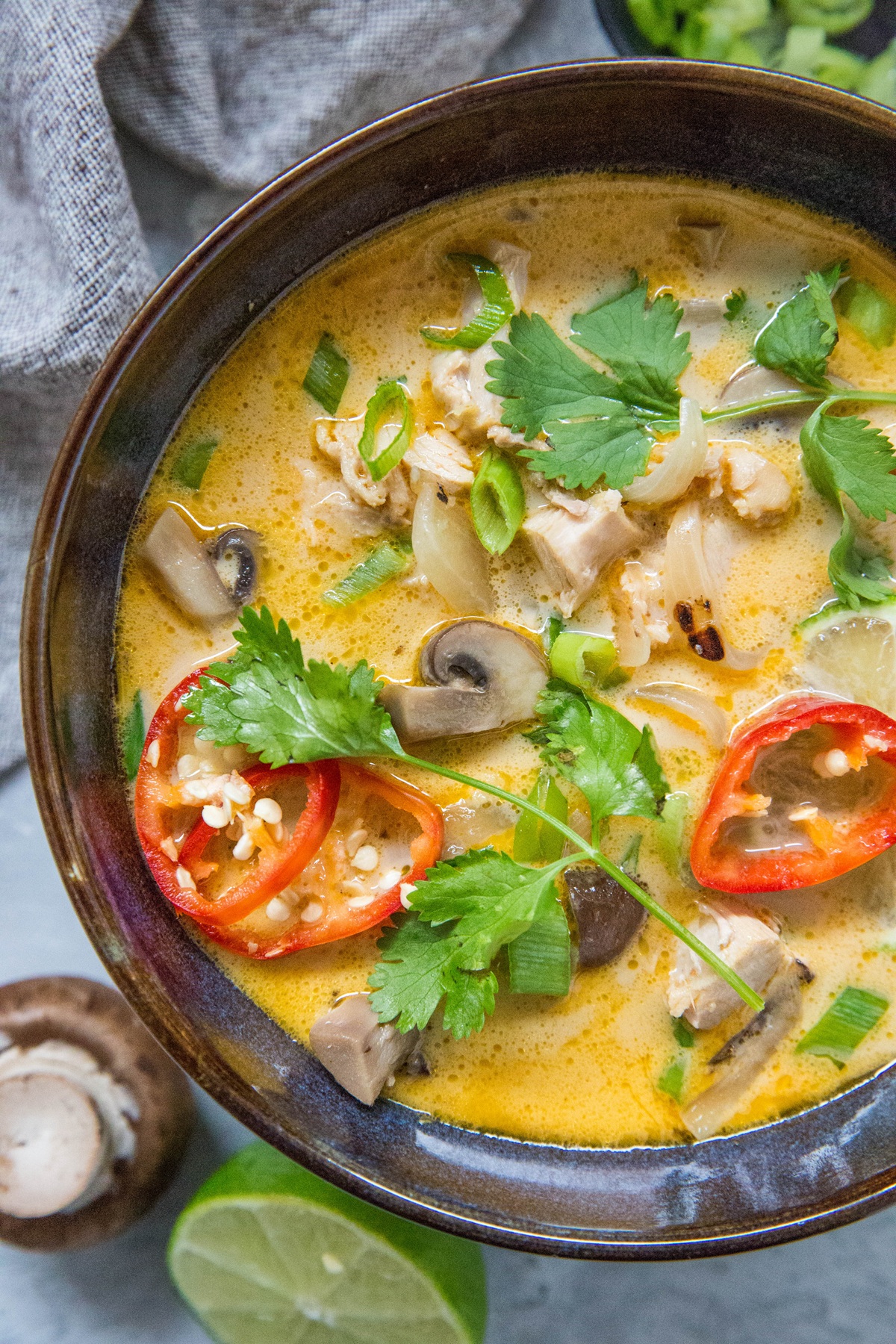 Blue ceramic bowl full of creamy Thai coconut chicken soup with mushrooms, cilantro, and red chilis on top. A fresh mushroom and half of a lime to the side.