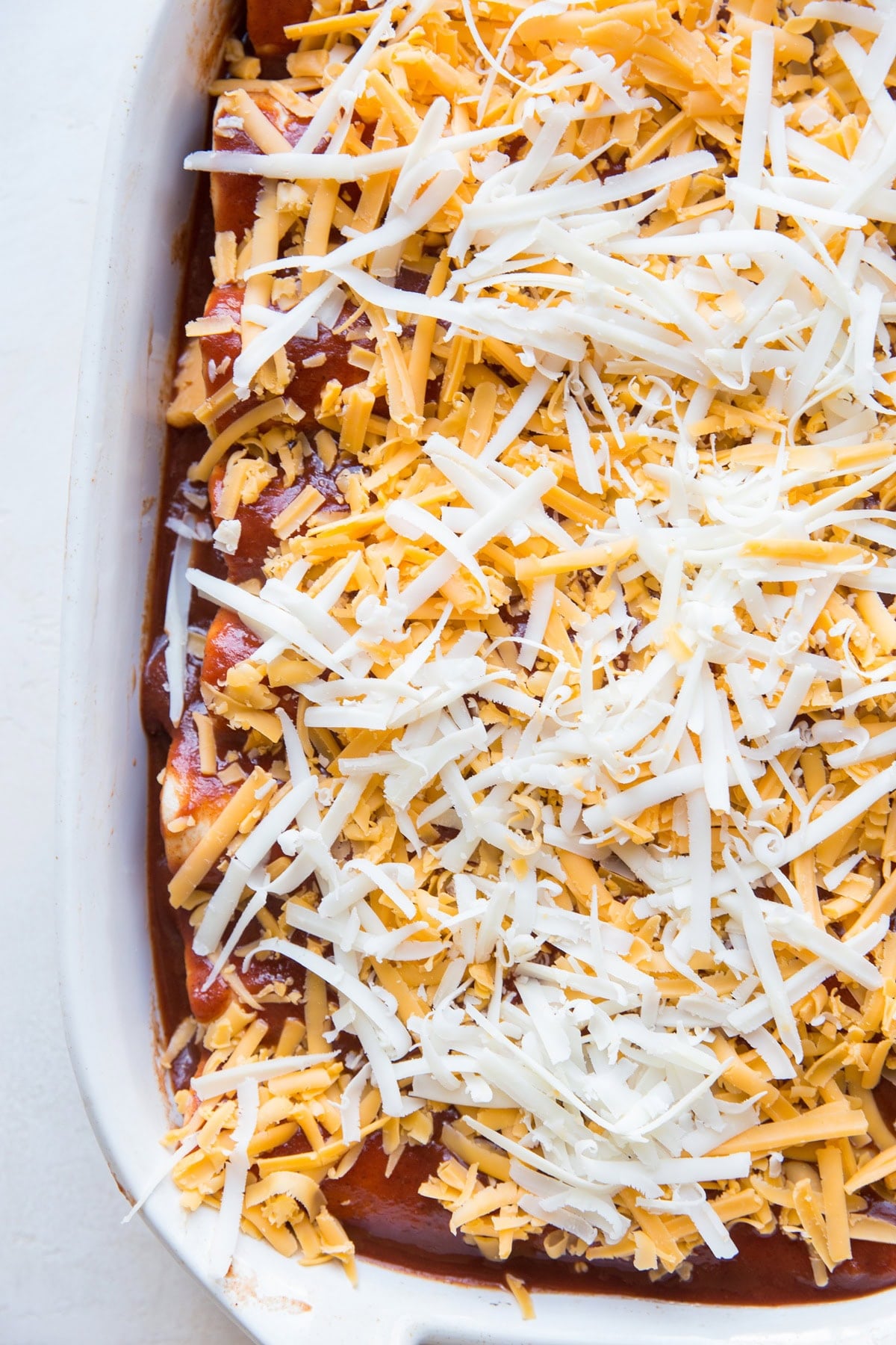 Pork enchiladas in a baking dish with enchilada sauce and a lot of grated cheese on top, ready to go into the oven.
