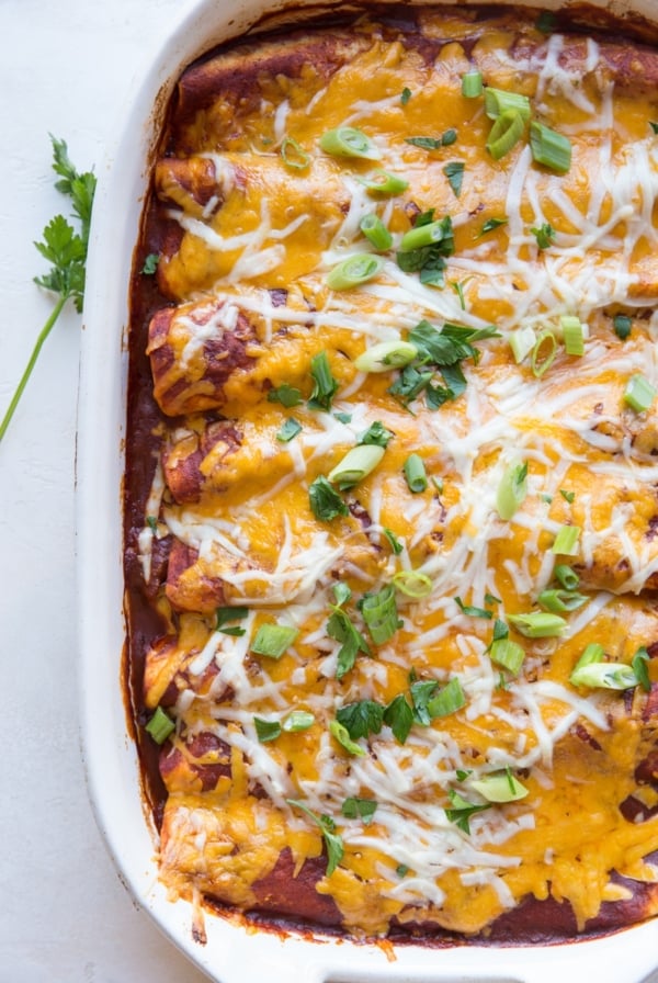 Casserole dish full of pork enchiladas with shredded cheese, chopped green onions, and parsley on top. Fresh out of the oven and ready to serve.