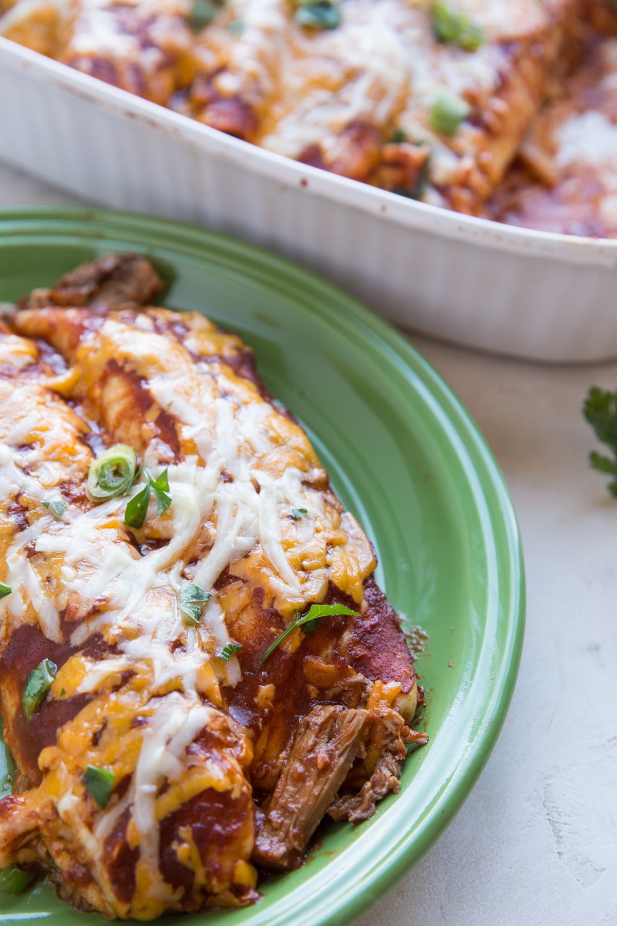 Green plate of shredded pork enchiladas with the rest of the pan of enchiladas in the background.