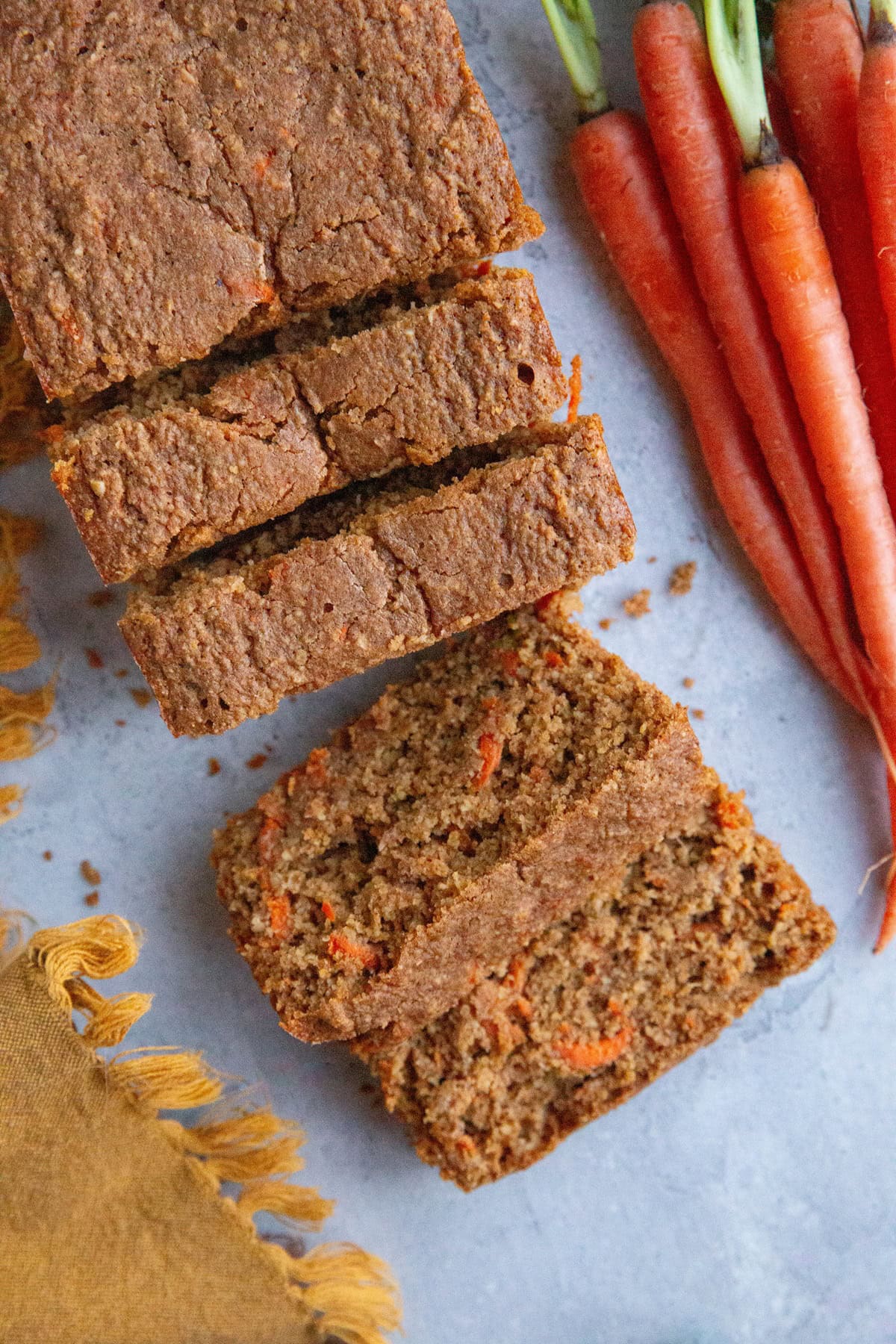 Carrot ginger cake cut into slices (in the form of a loaf) on a blue backdrop with fresh carrots and a golden napkin to the side. Ready to serve.