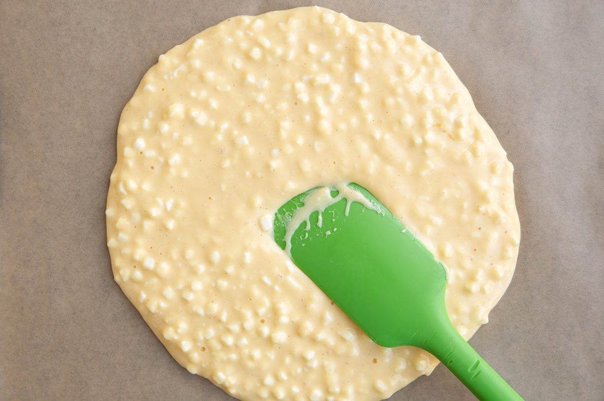 Rubber spatula spreading cottage cheese pizza crust mixture over a parchment-lined baking sheet.