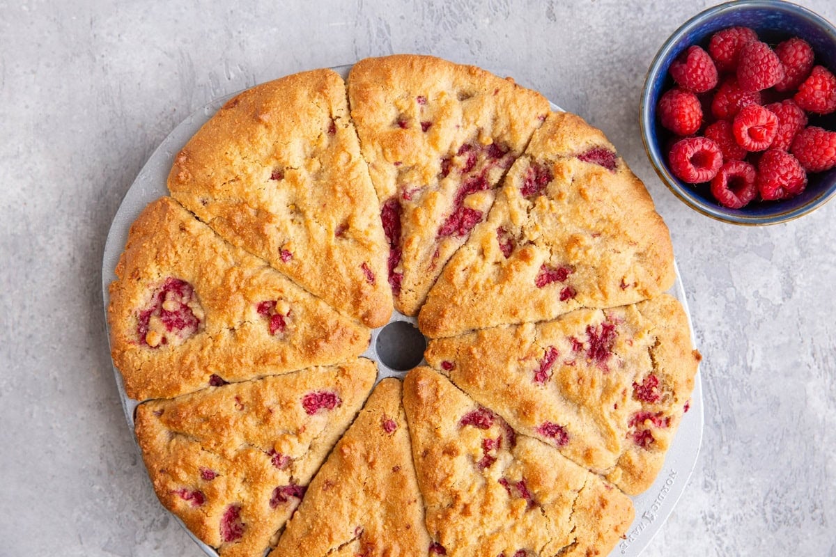 Scone pan filled with raspberry scones, fresh out of the oven, and a small blue bowl of raspberries to the side.