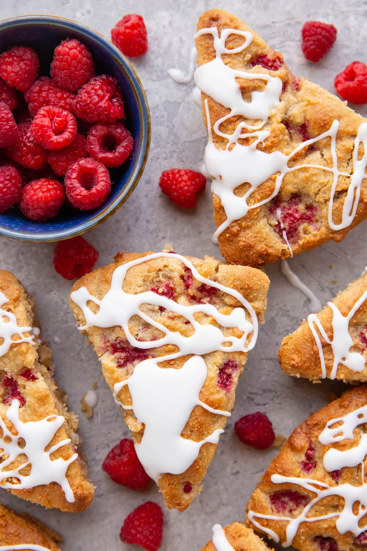 Six raspberry scones sitting on a background drizzled with glaze with fresh raspberries all around and a small bowl of raspberries to the side.