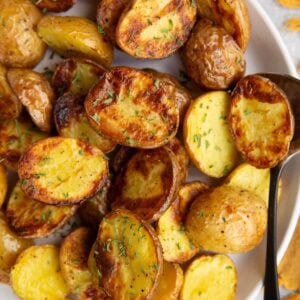 White plate full of golden brown baby potatoes that are sprinkled with dried parsley and a serving spoon is to the side.