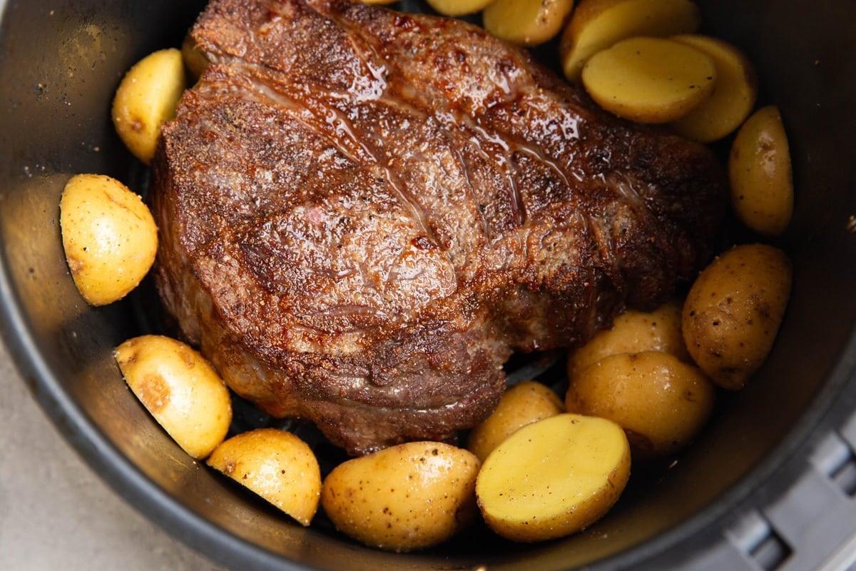 Basket of an air fryer full of chuck roast and raw potatoes that have just been added in to continue cooking.