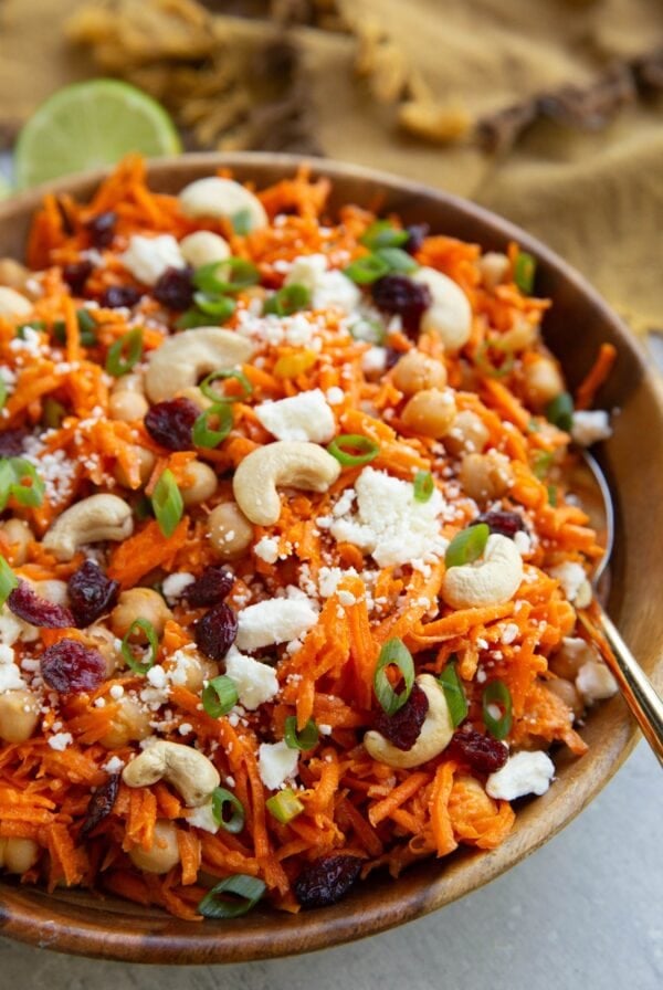 Wooden bowl of grated carrots, cashews, dried cranberries, chickpeas, and feta cheese with a golden spoon for serving and a fresh lime in the background.