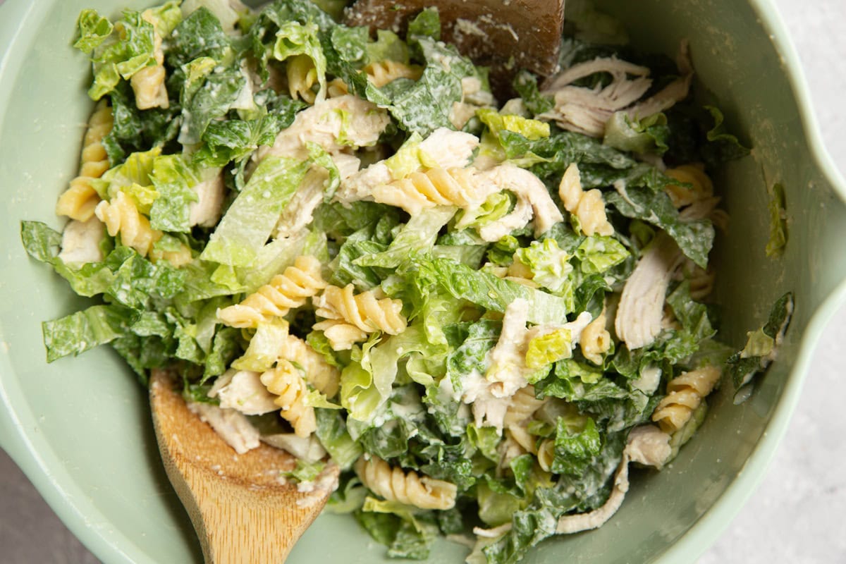 Mixing bowl with chicken Caesar pasta salad mixed up with two wooden spoons.