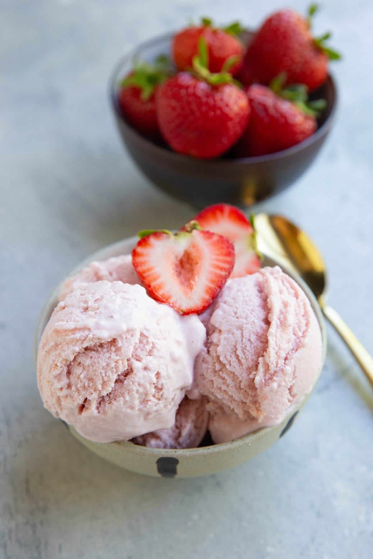 Bowl of strawberry cottage cheese ice cream with a golden spoon to the side and a bowl of strawberries in the background.