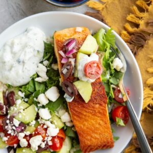 Big white bowl with brown rice, chopped lettuce, crispy seared salmon, Greek salad, feta cheese, and tzatziki sauce with a small blue bowl of more Greek salad to the side and a golden napkin.