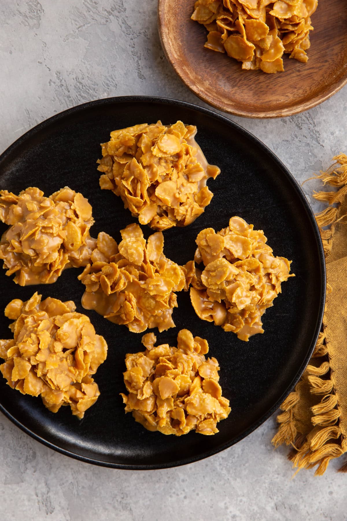 A black plate with six clusters of golden cornflake cookies on a gray surface, with a wooden bowl of loose cornflakes and a mustard-yellow fringed napkin nearby.