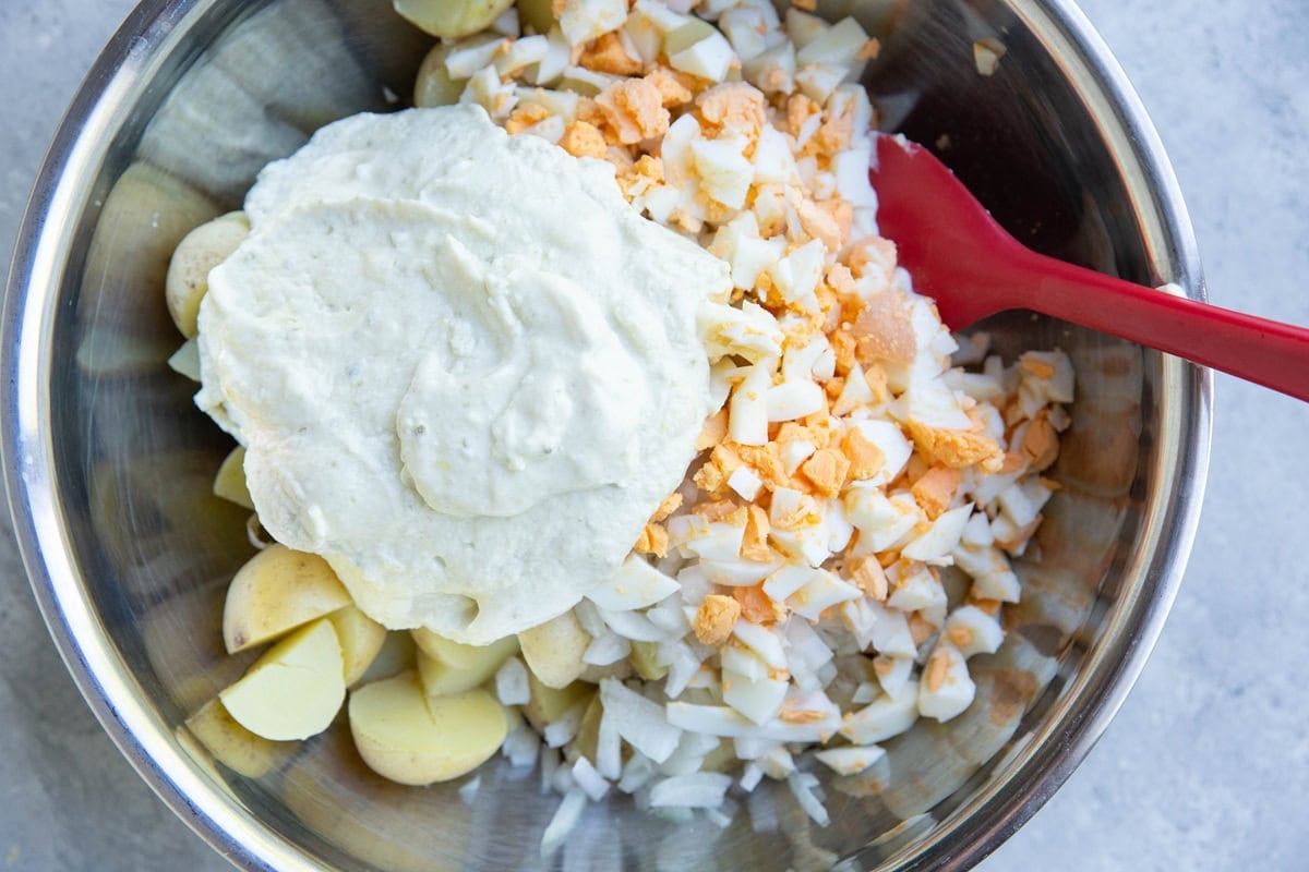 Stainless steel bowl full of chopped boiled potatoes, yellow onion, hard boiled eggs, and creamy cottage cheese dressing on top with a red rubber spatula to be mixed in.