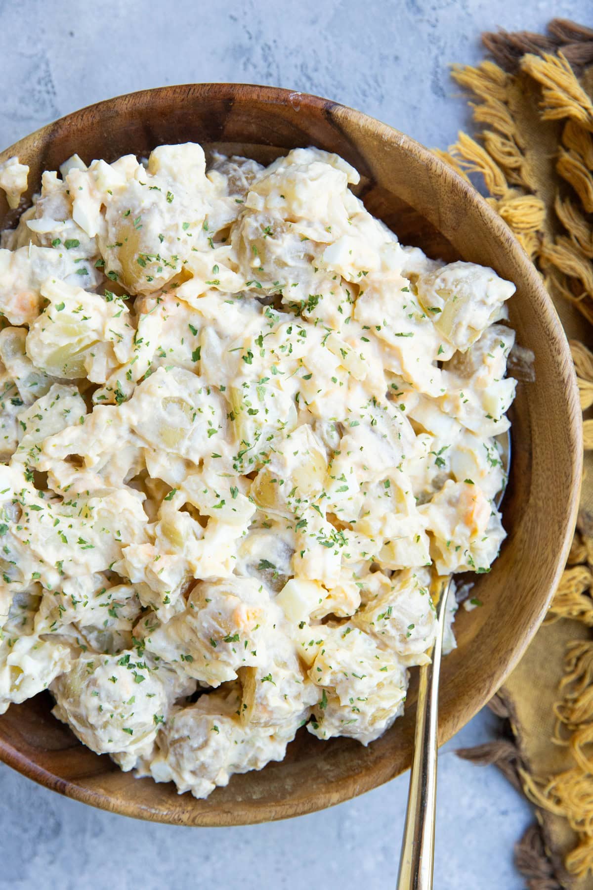 Cottage cheese potato salad in a large wooden bowl with dried parsley sprinkled on top, a gold spoon for serving and a gold napkin to the side.